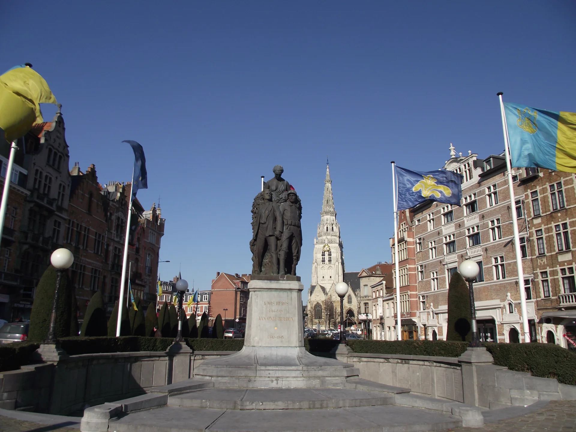 Statue et drapeaux au premier plan de la place de la Vaillance, avec les façades et la flèche de la collégiale en arrière-plan.