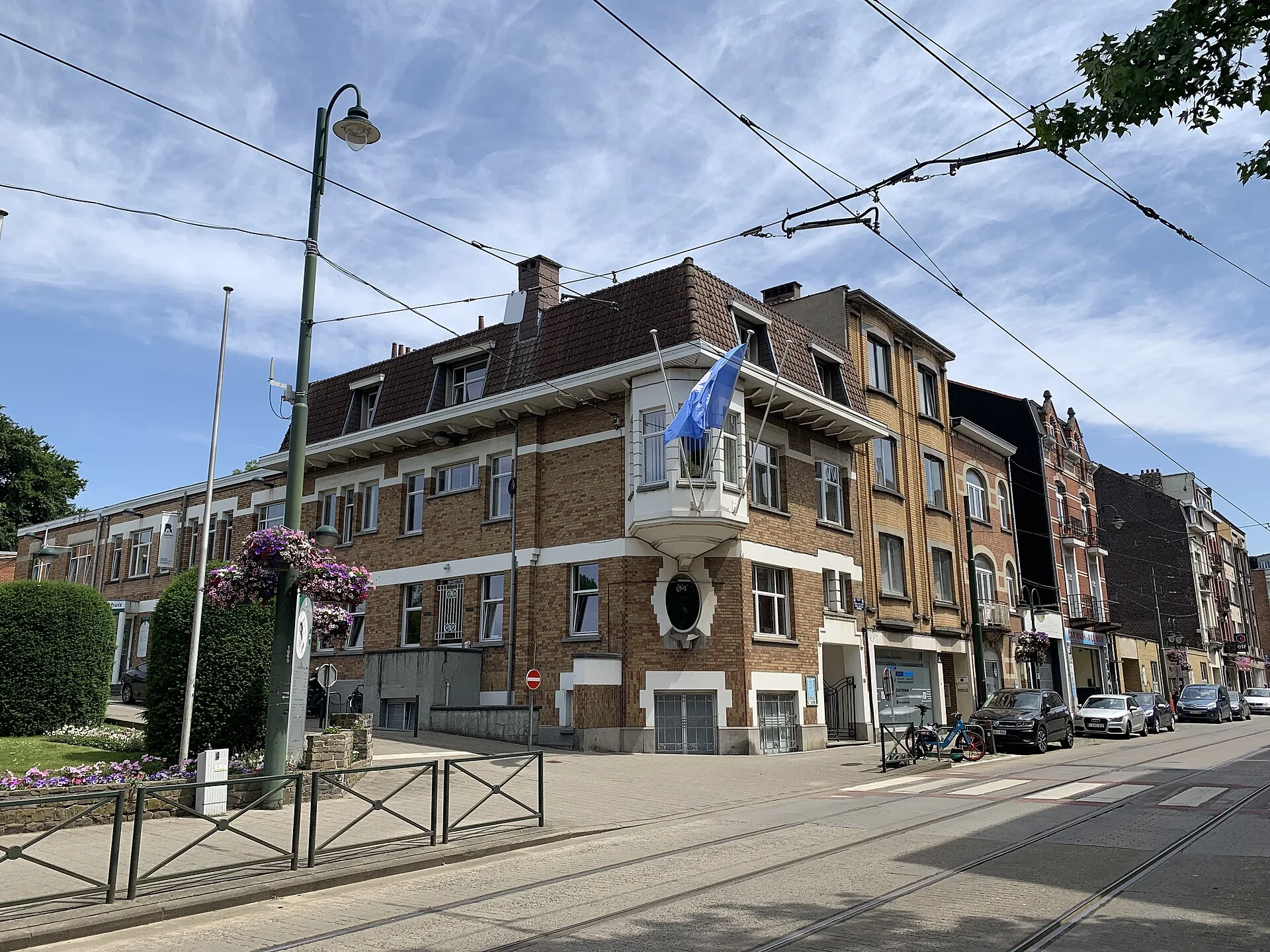 Maison communale en brique vue depuis la rue, avec les rails du tram au premier plan.