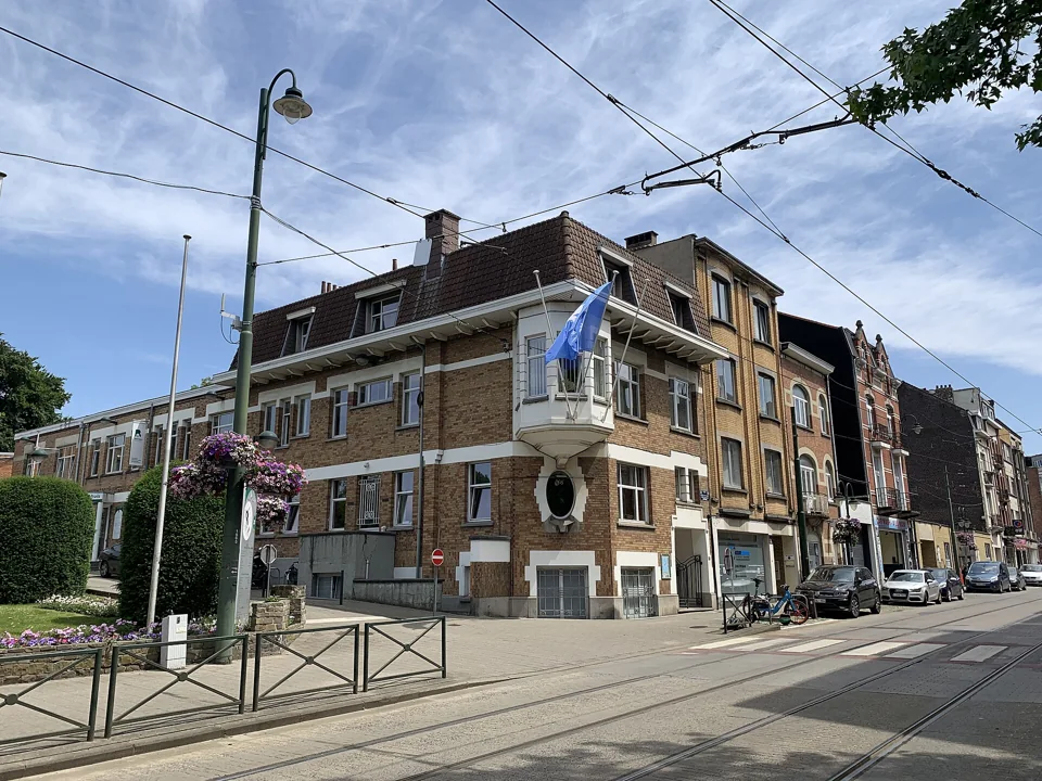 Maison communale en brique vue depuis la rue, avec les rails du tram au premier plan.