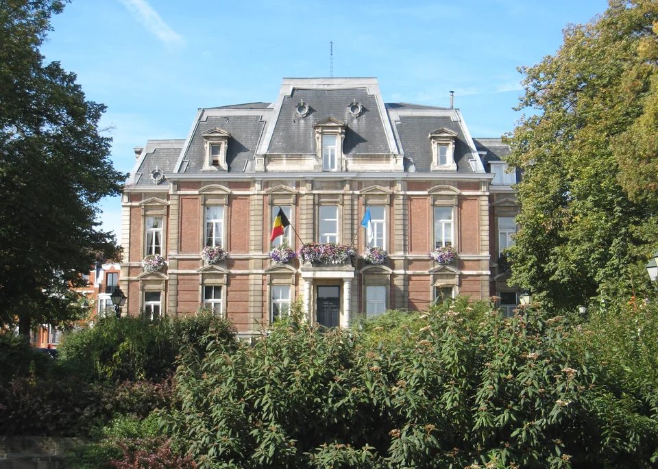Façade en brique et pierre de la maison communale d’Uccle, derrière une haie et des arbres.