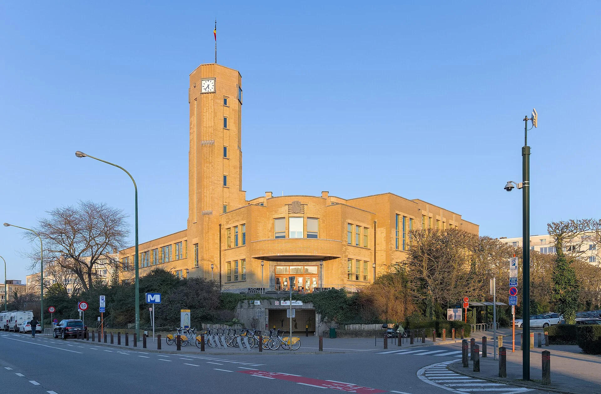 Maison communale en brique jaune, tour à horloge et avenue au premier plan.