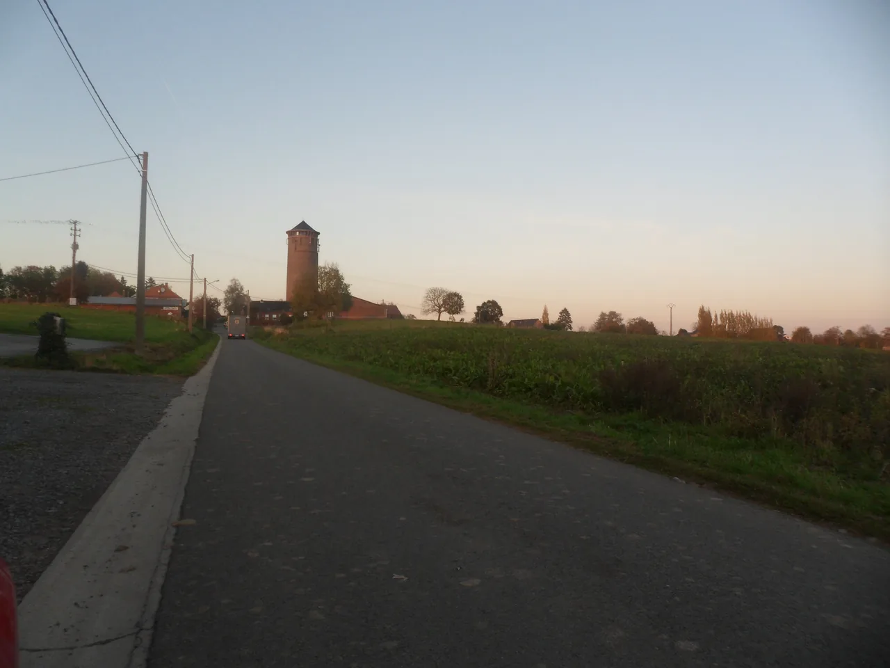 Route bordée de champs à Soignies, avec une tour en brique à l’arrière-plan.